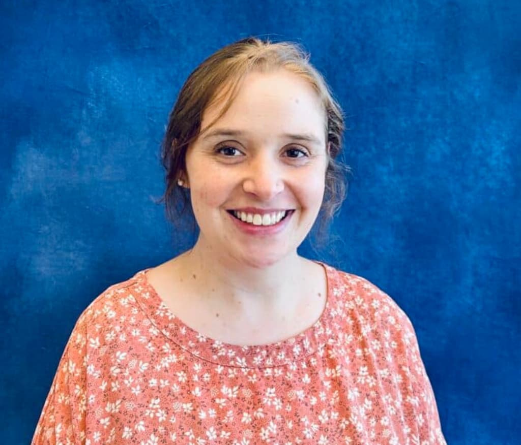 Smiling woman with long blonde hair, wearing a pink blouse, standing in front of a blue background.