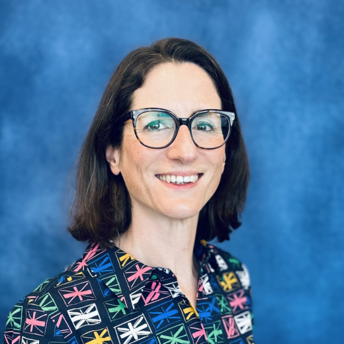 Smiling woman with short dark hair, wearing a colorful blouse, standing in front of a blue background.