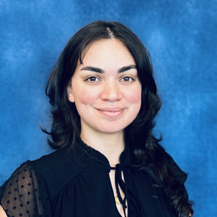 Alayna Self Smiling woman with long dark hair, wearing a black blouse with sheer dotted sleeves, standing in front of a blue background.