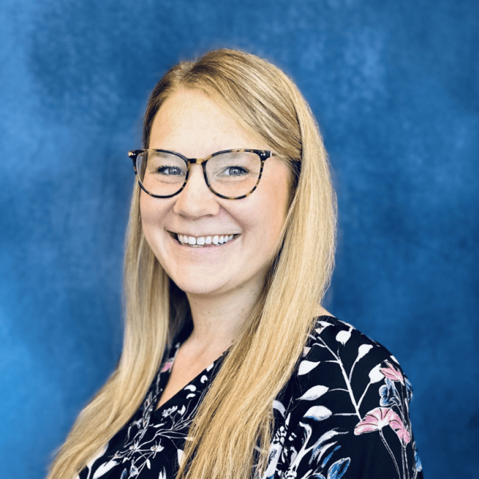 Smiling woman with long blonde hair, wearing glasses and a dark floral blouse, standing in front of a blue background.