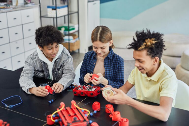 Three teens assembling a robotics kit with red components at a classroom table.