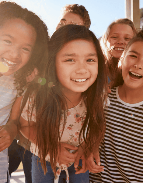 Group of smiling children standing close together outdoors, enjoying time in the sun.