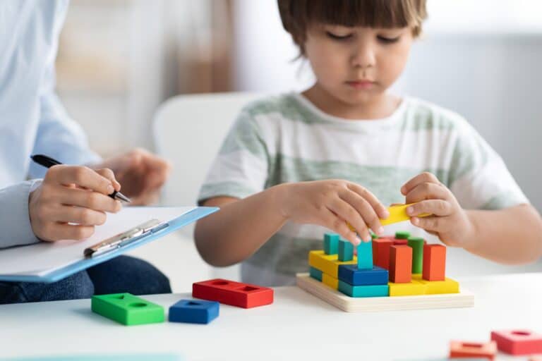 Child concentrating while stacking colorful wooden blocks, as an adult with a clipboard takes notes beside them.