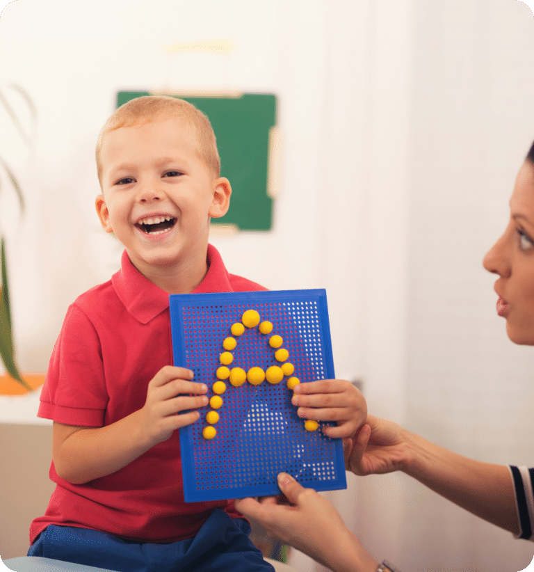 Smiling young boy in a red shirt holding a board with yellow pegs forming the letter A, while an adult assists beside him.