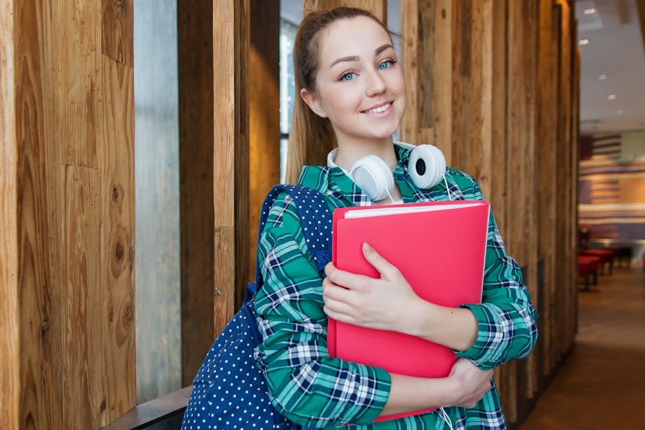 Smiling student with headphones and a polka-dot backpack holding a pink binder in a school hallway.